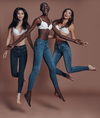 Wall Mural - So much fun! Full length of three attractive young women looking at camera and smiling while jumping against brown background