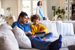 © Monkey Business - Close up of young father sitting on sofa in the living room with his pre-teen daughter using tablet computer, selective focus, mum in the background
