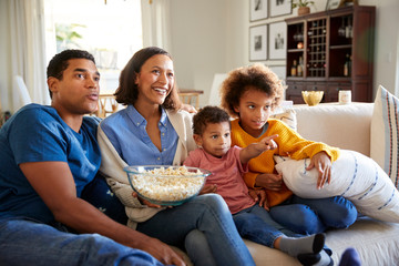 Wall Mural - Young family sitting together on the sofa in their living room watching TV and eating popcorn