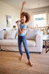 Wall Mural - Pre-teen girl dancing and singing in the living room at home using her phone as a microphone, full length, vertical