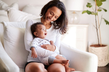 young adult mother sitting in an armchair in her bedroom, holding her three month old baby son in he