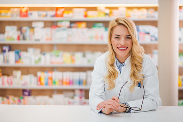 Wall Mural - Portrait of a cheerful young female pharmacist leaning on a counter at drugstore, looking at camera.