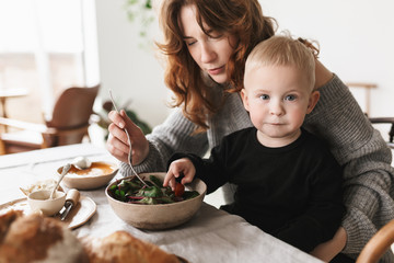 Young attractive mom with red hair in knitted sweater sitting at the table thoughtfully feeding her little son that dreamily looking in camera in kitchen at home