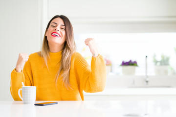 Young beautiful woman drinking a cup of coffee at home very happy and excited doing winner gesture with arms raised, smiling and screaming for success. Celebration concept.