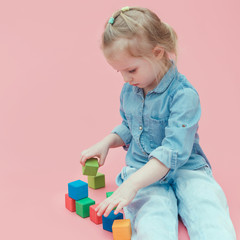 A charming little girl in denim clothes on a pink background plays with wooden colored cubes.