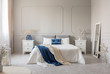 King size bed with grey, blue and white bedding between two wooden nightstands with flowers in vases, copy space on empty grey wall