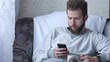 © smartbobert - One white young brunette curly-bearded man uses the phone sitting in a chair drinking tea or coffee