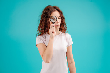 Wall Mural - Attractive brunette girl with curly hair isolated over blue turquoise background looking at something through magnifying glass with a big eye. Vision concept