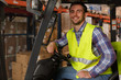 © Nestor - Forklift operator sitting in vehicle, posing and smiling. Handsome man wearing reflective coat while working in warehouse with transportation of goods and boxes.