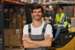 Front view of handsome workerof warehouse smiling, posing and looking at camera. Specialist wearing in acid green reflective waistcoat, loader operating forklift on background.