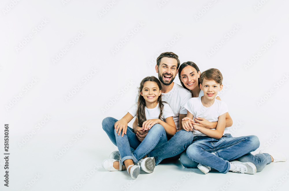 Relationship concept. Beautiful and happy smiling young family in white T-shirts are hugging and have a fun time together while sitting on the floor and looking on camera. - obrazy, fototapety, plakaty 