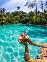 Cropped shot of girl holding coconut cocktail in outdoor swimming pool. Sunny day in tropical country.