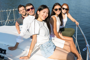 Positive international friends looking at camera, smiling and having fun together, wearing white clothes and sunglasses. Group of five friends sitting together on edge of boat bow and posing.