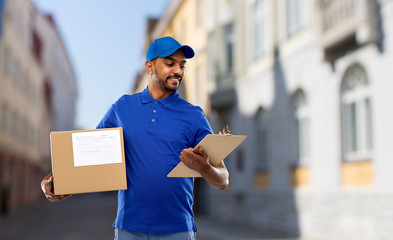 Wall Mural - mail service and shipment concept - happy indian delivery man with parcel box and clipboard in blue uniform over old tallinn city street background