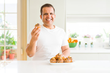 Middle age man eating chocolate chip muffins at home with a happy face standing and smiling with a confident smile showing teeth