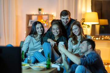 friendship, people, technology and entertainment concept - happy friends with smartphone, snacks and non-alcoholic drinks watching tv at home in evening