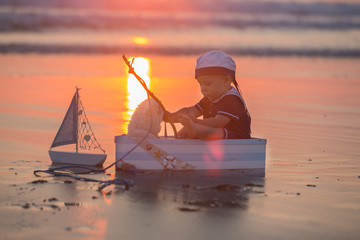 Wall Mural - Cute baby child, sweet boy, playing with boat, teddy bear and fishes on sunset at the edge of the ocean