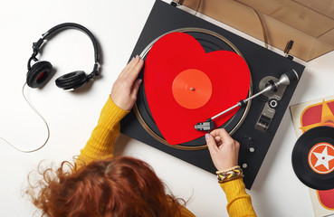 A red heart vinyl record turntable on a white table with plates. Included gramophone, black headphones. The hand of a girl DJ with bright accessory puts a stylus on a vinyl record. Valentine's Day