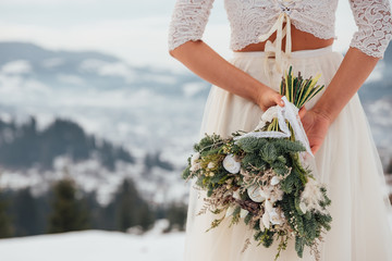 Bride in white wedding dress holding colorful flowers bouquet in hands and posing outdoors. Winter wedding and season floral concept.  Mountains on background.