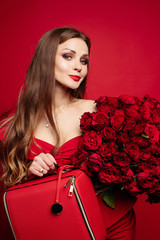 Wall Mural - Front view of gorgeous woman in red dress with makeup keeping beautiful bouquet of roses, looking at camera and smiling. Pretty model posing on red isolated background. Concept of valentines day.