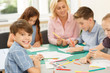 © Nestor - Pupils with teacher sitting at desk, enjoying painting with colorful pencil at school classroom and one boy smiling at camera. Girl explaining something to her classmate and woman helping children.