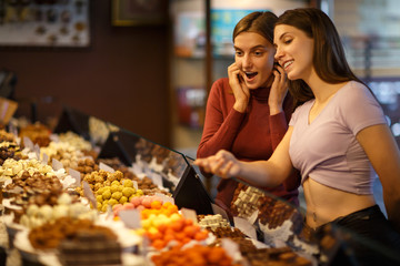 Two amazed girls standing next to showcase with assortment of candy.