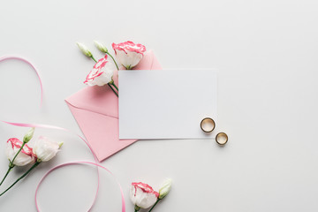 top view of empty card with pink envelope, flowers, silk ribbon and golden wedding rings on grey background