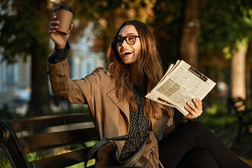 Sticker - Photo of happy woman drinking takeaway coffee and reading newspaper while sitting on bench in sunlit alley