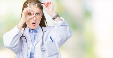 Poster - Middle age mature doctor woman wearing medical coat over isolated background doing ok gesture shocked with surprised face, eye looking through fingers. Unbelieving expression.