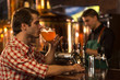 © Nestor - Side view of man holding glass and drinking delicious beer. Client of brewery wearing in checked shirt sitting at bar counter. Concentrated barmen in black shirt working behind.