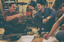 Friends From Rock Band Talking, Laughing And Composing Songs While Sitting On Floor. Guitarist In Checked Shirt Holding Electric Guitar. Singer Holding Professional Microphone, Wearing In Black Cap.