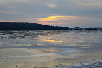 Wall Mural - The rays of the sunset sun breaking through the gray clouds and reflected in the surface of the lake with the remains of ice and dark banks covered with forest