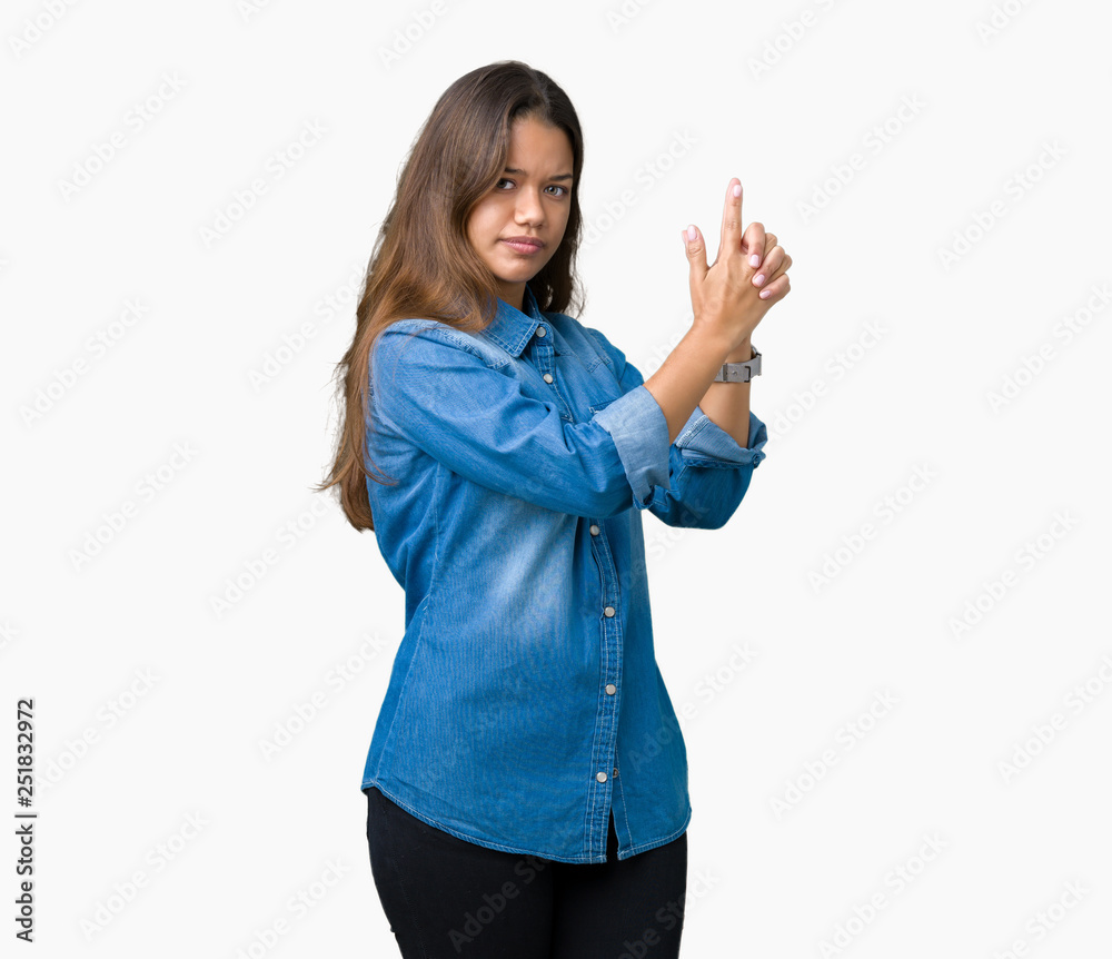 Young beautiful brunette woman wearing blue denim shirt over isolated background Holding symbolic gun with hand gesture, playing killing shooting weapons, angry face
