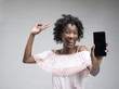 © master1305 - Portrait of a confident casual african american girl showing blank screen of mobile phone isolated over gray background at studio.