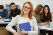 © Nestor - Portrait of young red haired girl with blue folder in hands. Happy beautiful student in glasses smiling in front of camera. Pupils sitting on lecture from foreign language in private school.