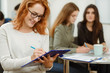 © Nestor - Red haired young student in glasses writing down new topic. Happy smiling girl in white sweater holding blue folder and smiling. Pupils on background feeling themselves comfortable during lessons.