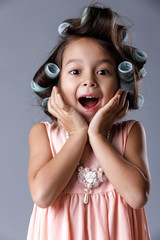 funny surprised happy little child girl in pink dress and hair curlers posing on gray background. hu