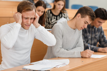 Confused male student doing difficult test and thinking over tasks during lecture at university. Young man worrying and keeping head with hands. Concept of anxiety and stressful situation.