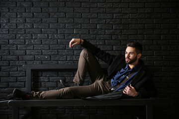 Wall Mural - Fashionable young man lying on bench against dark brick wall