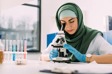 beautiful female muslim scientist looking through microscope during experiment in chemical laborator