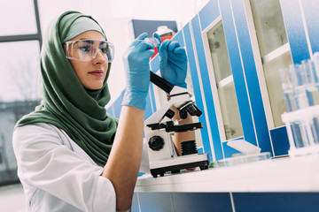 female muslim scientist sitting at table with microscope and looking at glass sample during experime