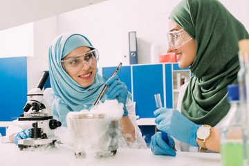 Wall Mural - smiling female muslim scientists experimenting with microscope and dry ice in chemical laboratory