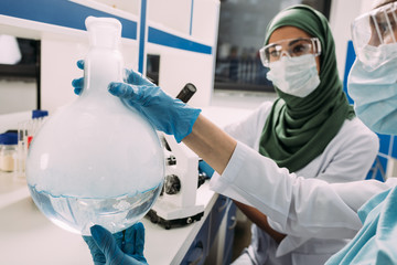 Wall Mural - female muslim scientists in goggles holding flask with liquid while experimenting in chemical laboratory