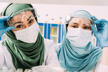 Wall Mural - female muslim chemists in medical masks and hijab looking at camera in laboratory