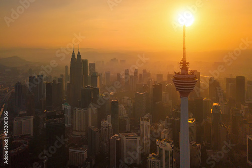 Plakat Menara Kuala Lumpur Tower ze słońcem. Widok z lotu ptaka centrum Kuala Lumpur, Malezja. Dzielnica finansowa i centra biznesowe w azjatyckim mieście miejskim. Wieżowiec i wieżowce o zachodzie słońca.