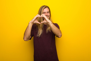 Wall Mural - Blond man with long hair over yellow wall making heart symbol by hands