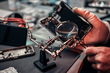 Wall Mural - Repairman uses magnifier and tweezers to repair damaged smartphone. Close-up photo of a disassembled smartphone.