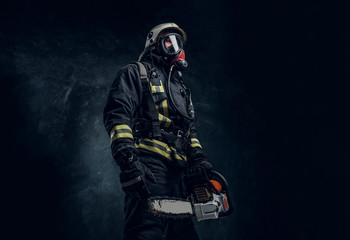 Wall Mural - Firefighter in safety helmet and oxygen mask holding a chainsaw. Studio photo against a dark textured wall