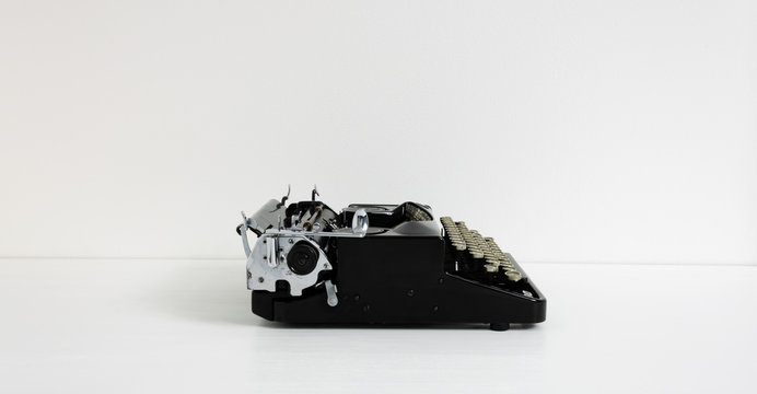 Old retro vintage black typewriter in black color laid on white desk in front of white wall with copy space