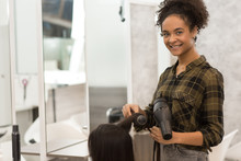 Young Happy Girl With Curly Hair Looking At Camera And Smiling. Professional Hair Cutter In Checked Shirt Working With Client In Beauty Salon. Hairdresser Holding Hair Dryer And Brush In Hands.